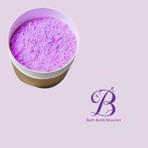 Purple bath bomb in a round open container against a lavender background, with the Bath Bomb Beauties logo visible nearby.