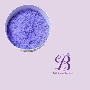 Purple bath bomb crumble in a round white tin on a pale lavender background, with the Bath Bomb Beauties logo in the bottom right corner.