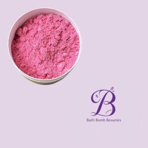 Pink bath bomb powder in a round container on a lavender background, with the Bath Bomb Beauties logo visible in the corner.