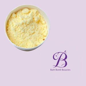 White tub filled with yellow crumbled bath powder on a lavender background, with Bath Bomb Beauties logo in the corner.