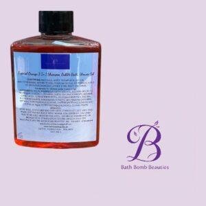Red-orange bottle with black cap containing 3-in-1 shampoo, bubble bath and shower gel, label visible, beside Bath Bomb Beauties logo on a pale purple background.