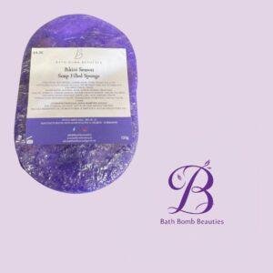 Purple oval soap package labeled 'Bikini Season Soap Filled Sponge' from Bath Bomb Beauties, with a purple logo and text in the bottom-right corner.