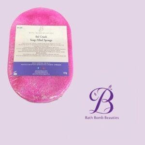 Pink soap filled sponge bath bomb labeled sol crush from bath bomb beauties placed on a lavender background with a decorative bath bomb beauties logo in the corner