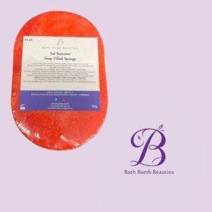 Red oval soap bar in plastic wrapper labeled sol summer soap filled sponge beside a bath bomb beauties logo on a lavender background