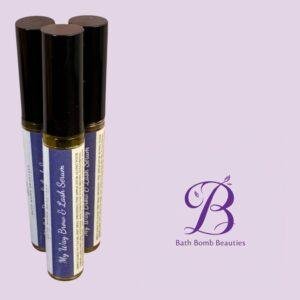 Three blue cylindrical bottles with black caps standing on a lavender background, featuring a Bath Bomb Beauties logo in the corner.