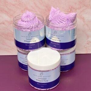 Four lavender whipped soap jars labeled Bath Bomb Beauties, Berry Comfort, arranged on a pink countertop with a white lid in front. The soap swirls peek from the open jars in a soft purple color.