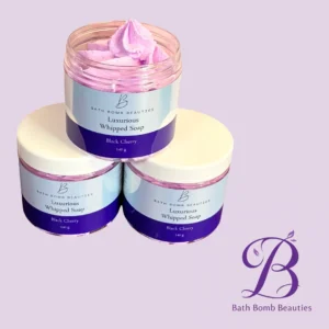 Three jars of Bath Bomb Beauties Luxurious Whipped Soap in Black Cherry with pink whipped soap on top, set against a lavender background with the brand logo in the corner.