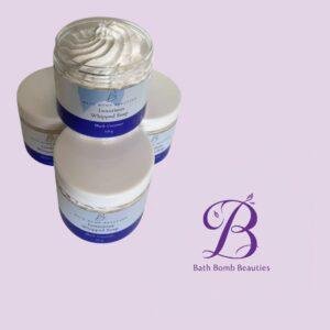 Stack of white jars with blue labels reading luxurious whipped soap and black coconut on a lavender background with bath bomb beauties logo in purple at the bottom right