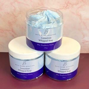 Three jars of Bath Bomb Beauties Luxurious Whipped Soap in Blue Raspberry Slushy scent, stacked with one on top of two, on a pink surface.