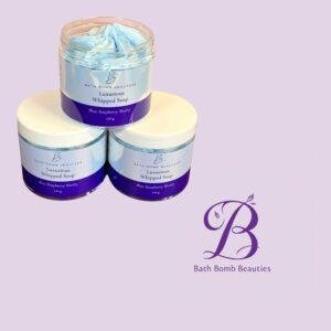 Jar of blue whipped soap from Bath Bomb Beauties, labeled Luxurious Whipped Soap, Blue Raspberry Slushy, stacked on a white surface against a pastel lilac background.