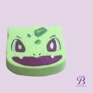 Green bath bomb shaped like a cute, mischievous face with purple eyes and a wide smile on a lavender background (Bath Bomb Beauties).