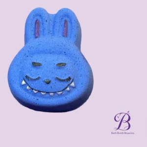 Blue bath bomb shaped like a cute creature with a smiling face on a lavender background bath bomb beauties logo in the corner