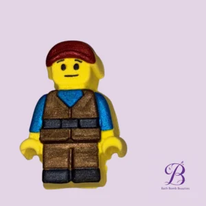 Yellow playmobil style figurine with red hair brown vest and blue sleeves against a lavender background bath bomb beauties logo at bottom right