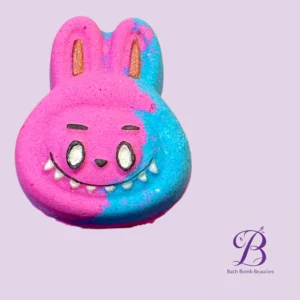 Two-tone bunny-shaped bath bomb (pink left, blue right) with a smiling face on a lavender background, Bath Bomb Beauties logo in the corner.