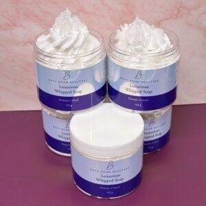 Four jars of Bath Bomb Beauties Luxurious Whipped Soap in Dream Cloud scent, topped with whipped cream, on a pink marble backdrop (foam visible).