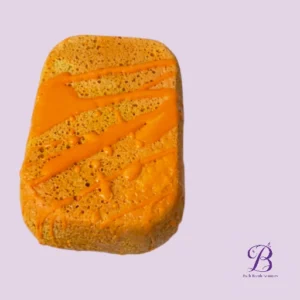 Orange frosted cake slice with orange drizzle on a light purple background, logo bottom right.