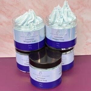 Four jars of Luxurious Whipped Soap by Bath Bomb Beauties, light blue whipped soap in clear jars on a pink surface.