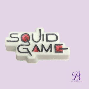 White bath bomb shaped like a game block with squid game lettering in black and red on a lavender background bath bomb beauties logo in the corner
