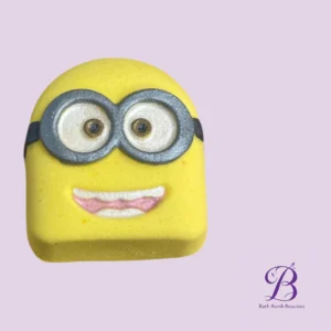 Yellow Minion-themed bath bomb with gray goggles and a pink-smiled mouth on a lilac background; Bath Bomb Beauties logo in the corner.