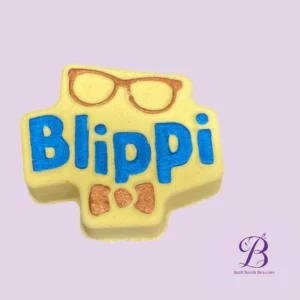 Yellow cross-shaped bath bomb with blue 'Blippi' lettering and orange glasses and bowtie details; Bath Bomb Beauties logo in the corner on a pale purple background.