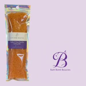 Holographic, sealed pouch of orange bath salts labeled “Peaches & Cream Dead Sea Soaking Salts” from Bath Bomb Beauties against a lavender background.