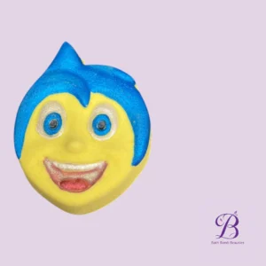 Face-shaped yellow bath bomb with blue hair and a wide smile on a light purple background; Bath Bomb Beauties logo in the corner.