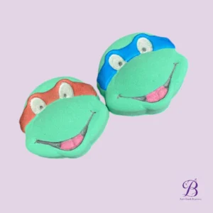 Two green frog-shaped bath bombs with red and blue bands, on a lavender background.