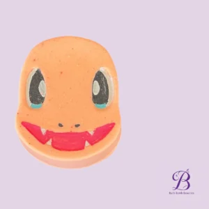 Orange smiley-face bath bomb with large black eyes and a red toothy grin on a light purple background; Bath Bomb Beauties logo in the bottom-right corner.