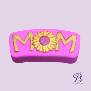 Bright pink soap bar engraved with 'MOM' in gold glitter letters and a sunflower in the center.