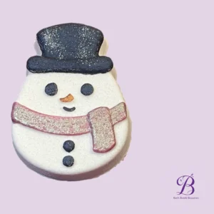 Snowman-shaped cookie with black hat, coal eyes, carrot nose, and pink scarf on a purple background; Bath Bomb Beauties logo visible in corner.