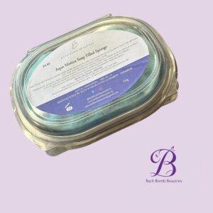 Close-up of a glass jar with a white label reading 'Aqua Mistica Soap Filled Sponge' from Bath Bomb Beauties, set against a lavender background.