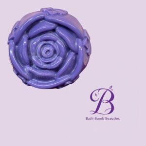 Close-up of a purple, swirled bath bomb against a pale lavender background with 'Bath Bomb Beauties' logo partially visible.