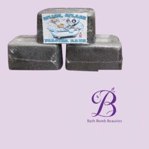 Three black rectangular bath bombs stacked on a lavender background with a playful label showing a cartoon bathtub edge; Bath Bomb Beauties logo in the corner.