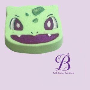 Green cartoon-faced bath bomb with purple eyes and a wide purple smile on a light purple background; Bath Bomb Beauties logo in the lower-right corner.