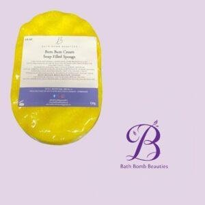 Bright yellow soap filled sponge with a white label reading bum bum cream soap filled sponge from bath bomb beauties on a lavender background with purple logo