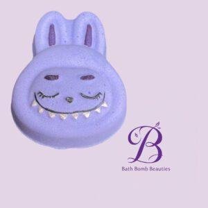Purple bunny shaped bath bomb with a smiling face on a lavender background features bath bomb beauties logo in the corner
