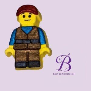 Lego style yellow minifigure with brown vest and blue sleeves on a pale purple background bath bomb beauties logo visible