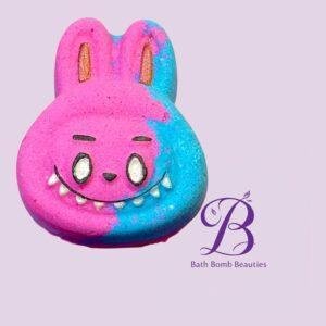 Colorful bunny-shaped bath bomb, pink and blue, with gold foil eyes, on a lavender background with the Bath Bomb Beauties logo nearby.
