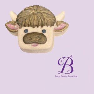 Cow shaped bath bomb with gold accents and pink ears on a lavender background featuring bath bomb beauties logo