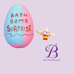 Pastel blue pink bath bomb with bath bomb surprise text beside a small cartoon bunny on a lavender background with bath bomb beauties logo nearby