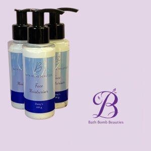Three Bath Bomb Beauties face moisturiser bottles with pump tops, blue labels, arranged against a lavender background with the brand logo nearby, 100 g each