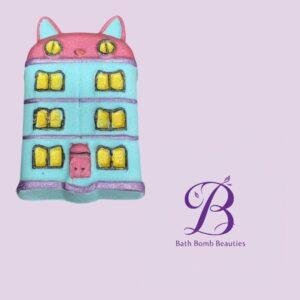 Pastel cat shaped bath bomb in blue and pink with yellow windows set against a lavender background with bath bomb beauties logo
