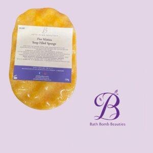 Orange flor mistica soap filled sponge bath bomb with label from bath bomb beauties placed on a lavender background with a purple logo nearby