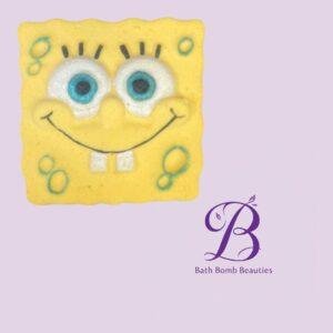 SpongeBob-like yellow square bath bomb with a smiling cartoon face, on a lavender background with the Bath Bomb Beauties logo.