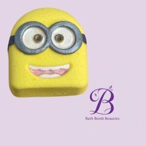 Yellow Minion-shaped bath bomb with gray goggles and a pink smile on a lavender background; Bath Bomb Beauties logo in the corner.