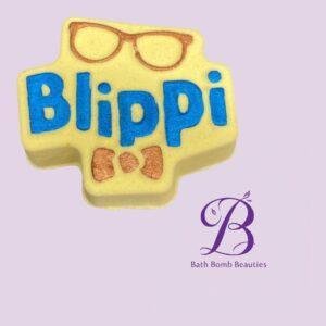 Blippi themed yellow cross shaped bath bomb with blue blippi lettering and copper glasses on a lavender background bath bomb beauties logo shown