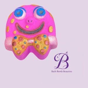 Pink smiley faced bath bomb with a gold polka dotted bow on a light purple background bath bomb beauties logo appears nearby