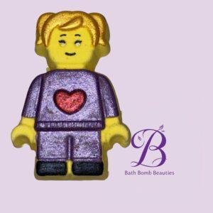 Lego style yellow minifigure in a purple outfit with a red heart on the chest set against a lavender background with a bath bomb beauties logo