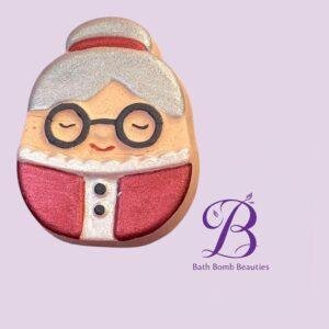 A smiley gingerbread cookie with round glasses and gray hair wearing a red outfit, against a lavender background with a Bath Bomb Beauties logo.