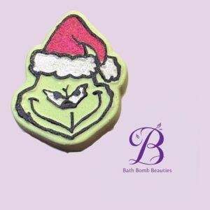 Green smiley-face bath bomb wearing a red Santa hat with white trim on a pale lavender background; Bath Bomb Beauties logo in purple at the bottom right.